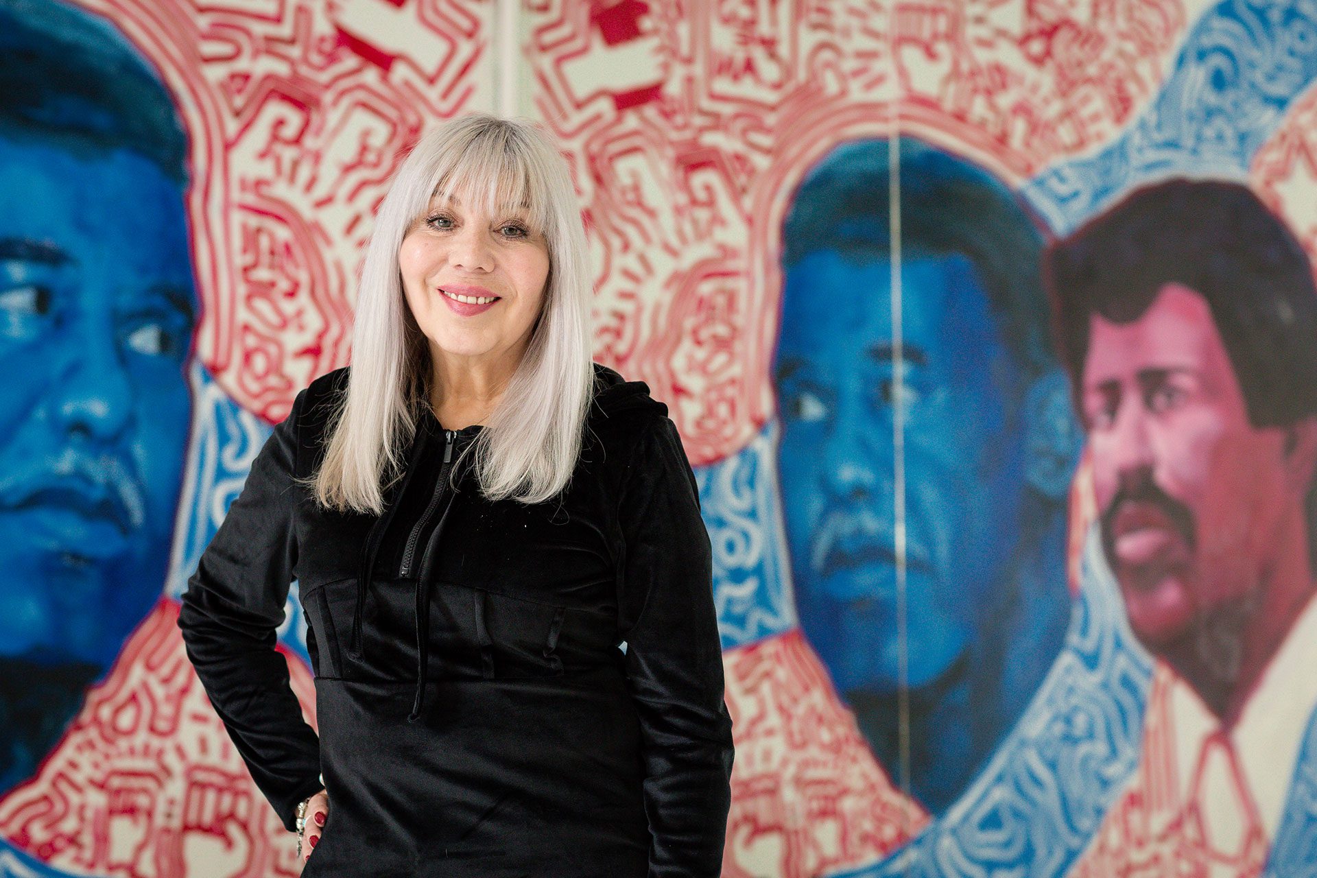 La organizadora y pastora Emma Lozano, vestida con una sudadera negra, posa delante de un mural que representa al alcalde de Chicago Harold Washington y al hermano de Lozano, Rudy Lozano.