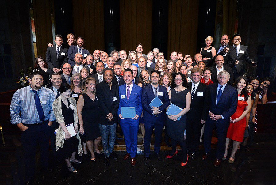 A group of about 50 on a stage with Viet Thanh Nguyen in the center in a blue suit receiving the Pulitzer Prize