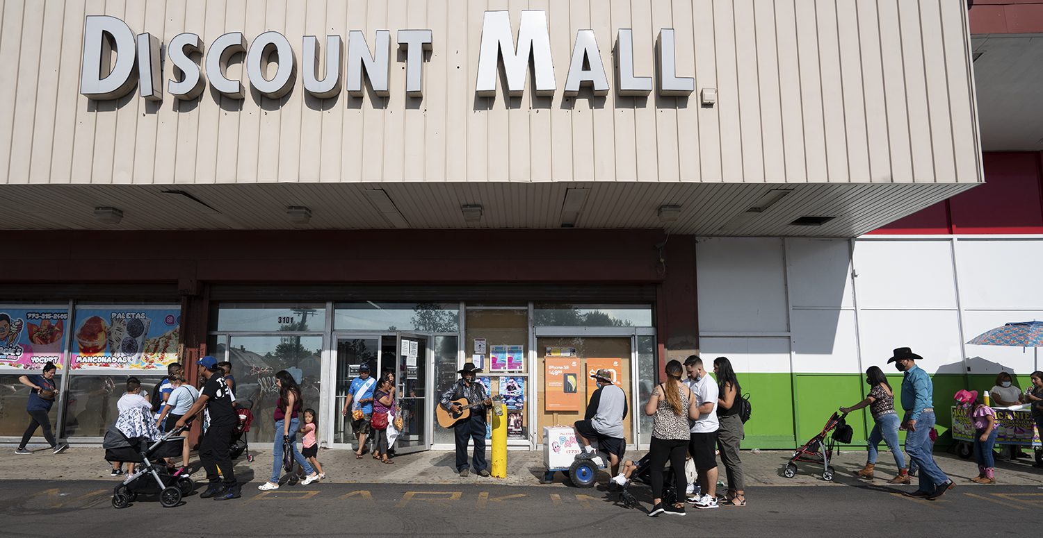 La gente camina hacia el centro comercial de descuentos de Little Village, en el centro un hombre toca la guitarra; hay varias personas con cochecitos