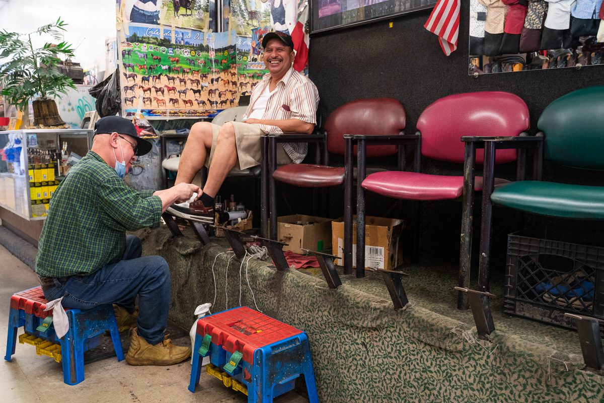 Mario Santo polishes the shoes of Andres Juarez who sits on the end of a row of chairs smiling