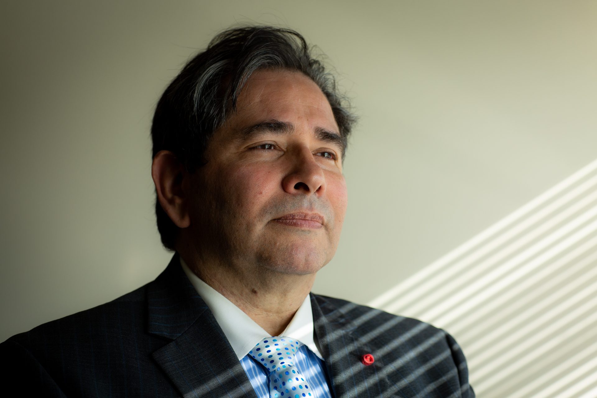 Frank Sandoval wearing a gray jacket and blue dot tie in front of a cream wall with rays of sunlight