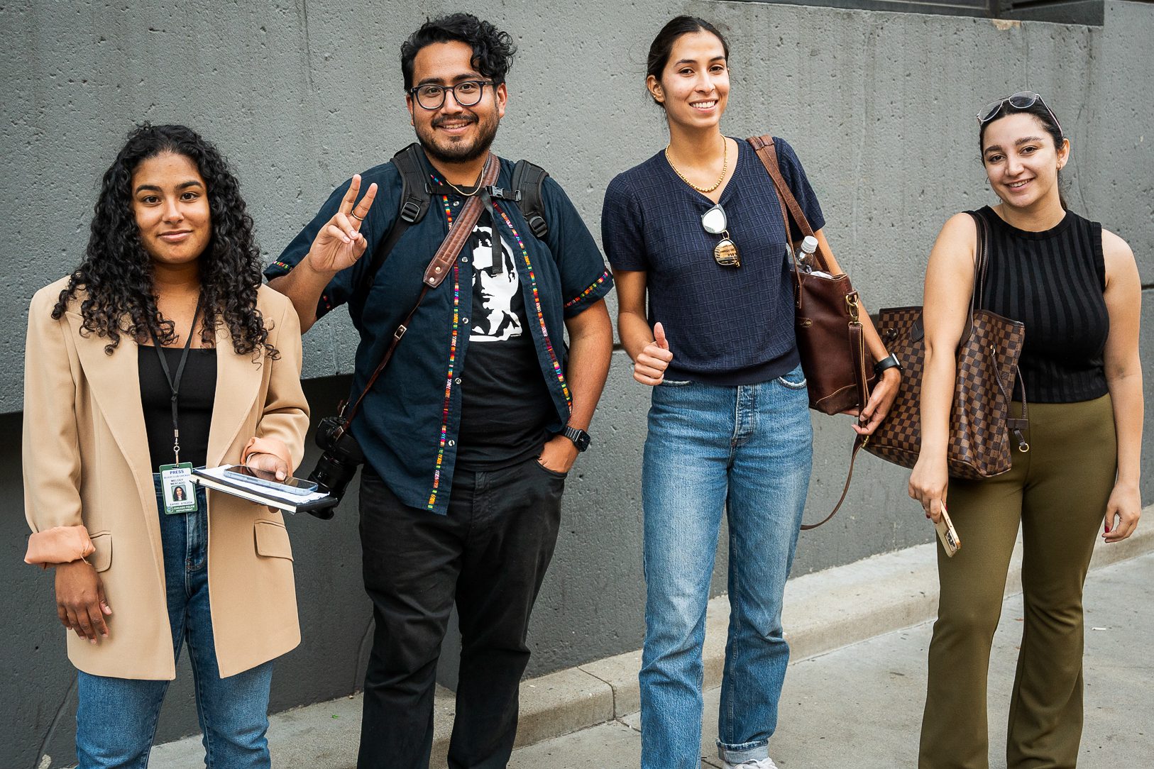 cuatro reporteros delante de un muro de cemento gris: Melody con un blazer beige Jesús con vaqueros negros un t-shrit negro y una chaqueta, Madison dando un pulgar hacia arriba y Diane con gafas en la cabeza.