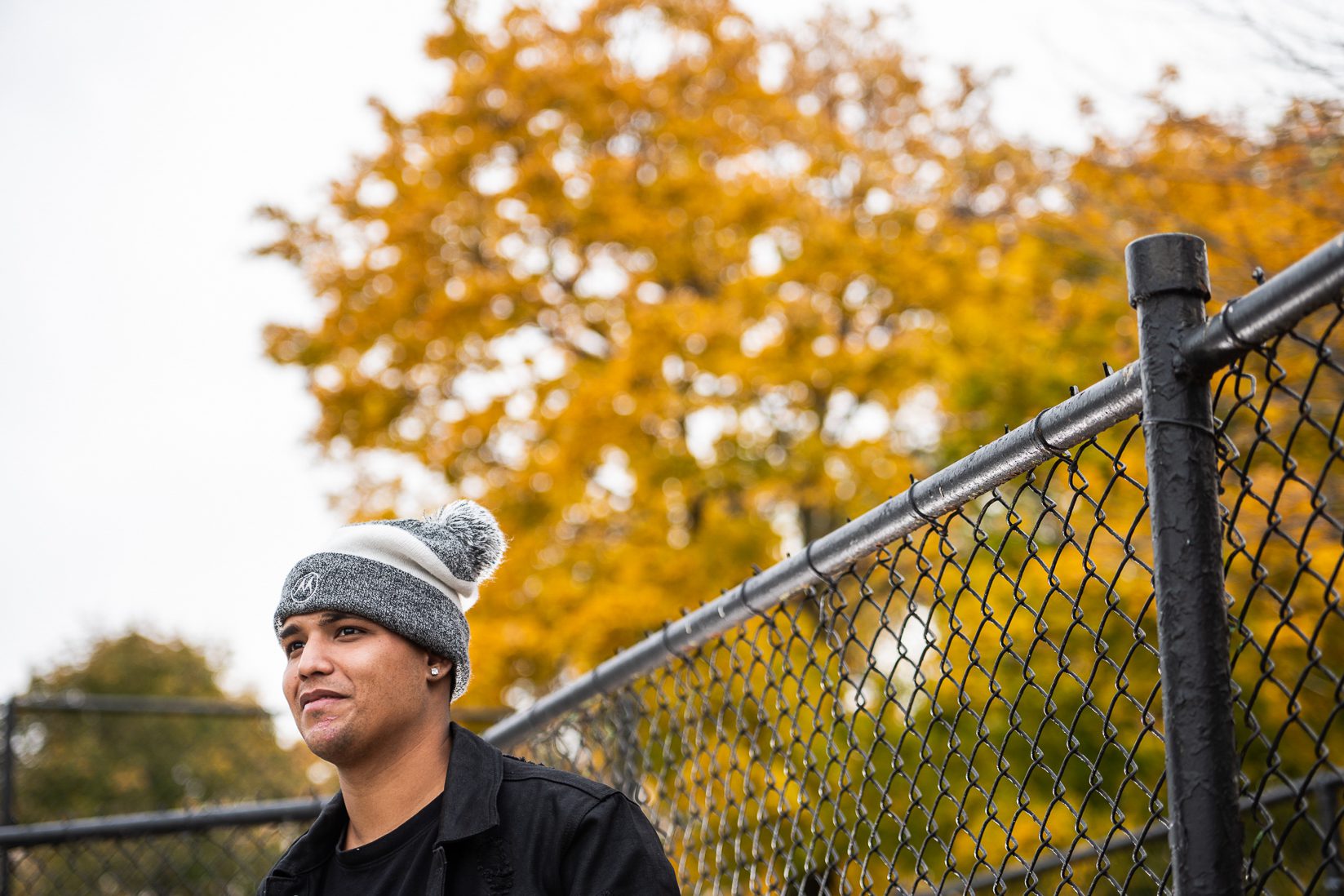 Nolram wears a gray and white beanie with a black shirt and jacket and black fence behind him and behind that yellow fall leaves