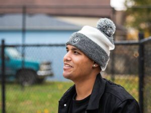 Nolram wears a gray and white beanie with a black shirt and jacket and black fence behind him