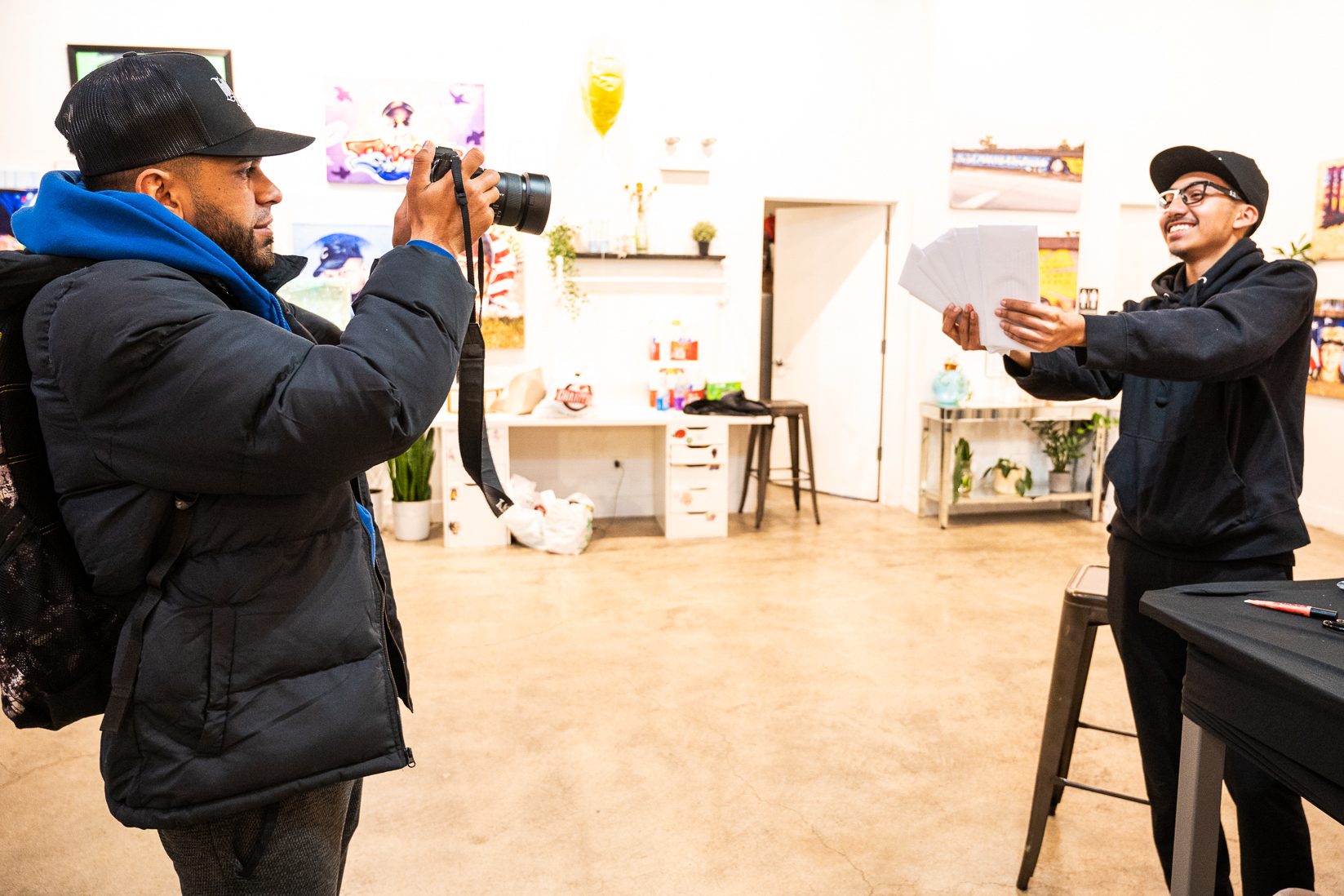 Jesus David Lucena wearing a black puffy coat and cap takes a photo of The Kid wearing all black and glasses with one side white and one black holding up white cards