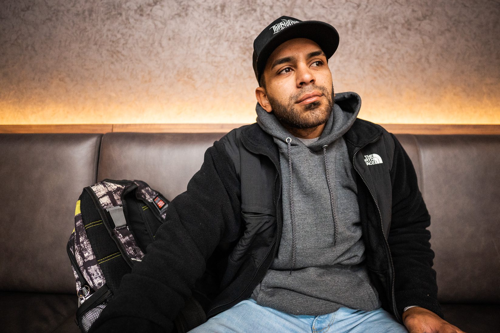 Jesus David Lucena Castillo sitting on a booth bench wearing a gray hoodie and black jacket with a cap and backpack