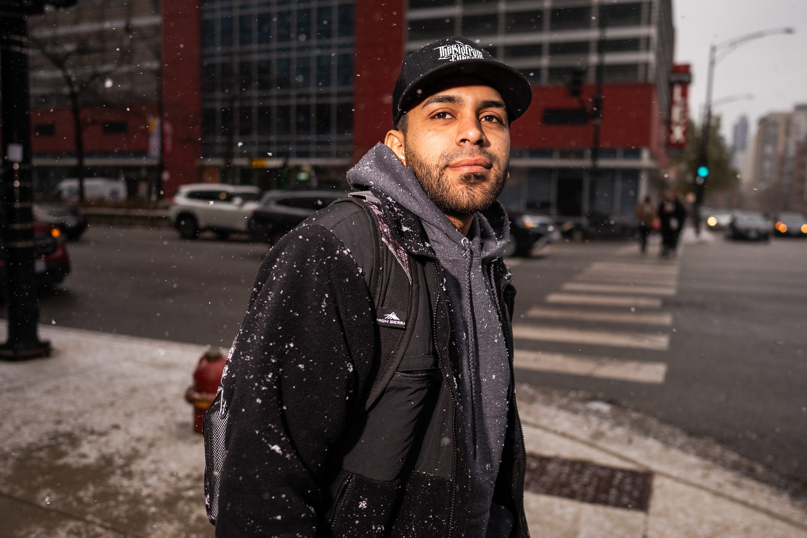 Jesus David Lucena Castillo a gray hoodie and black jacket standing outside on the sidewalk in Chicago as the snow falls down