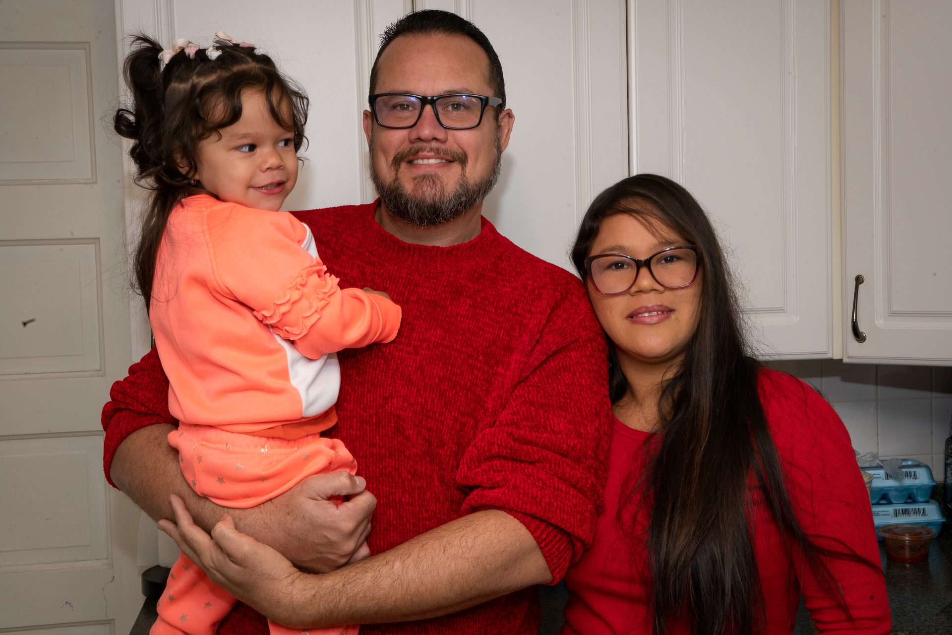 Tomas and Carmen wear red sweater in their white kitchen, their daughter Grecia is in organe pajams