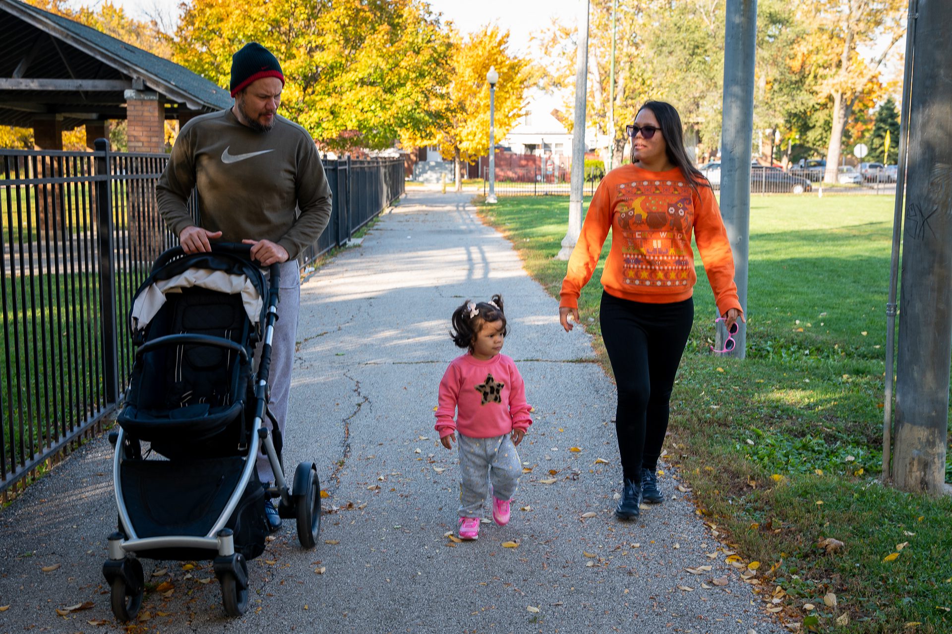 Tomas pushes the stroller while Carmen and Grecia walk, they are all on the path by the park