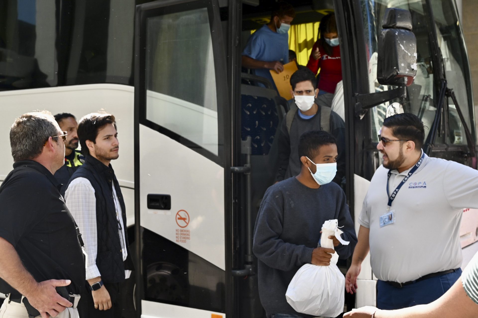 inmigrantes bajando de un autobús con máscaras y guardando sus pertenencias en bolsas de plástico