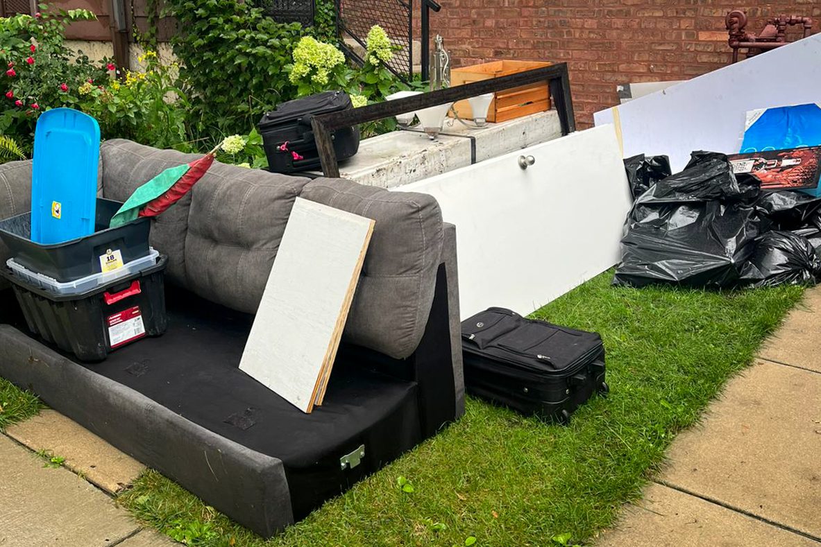 un sofá con los cojines arrancados, cajas, maletas, luces, una puerta y bolsas de basura se sientan en un pequeño jardín delantero