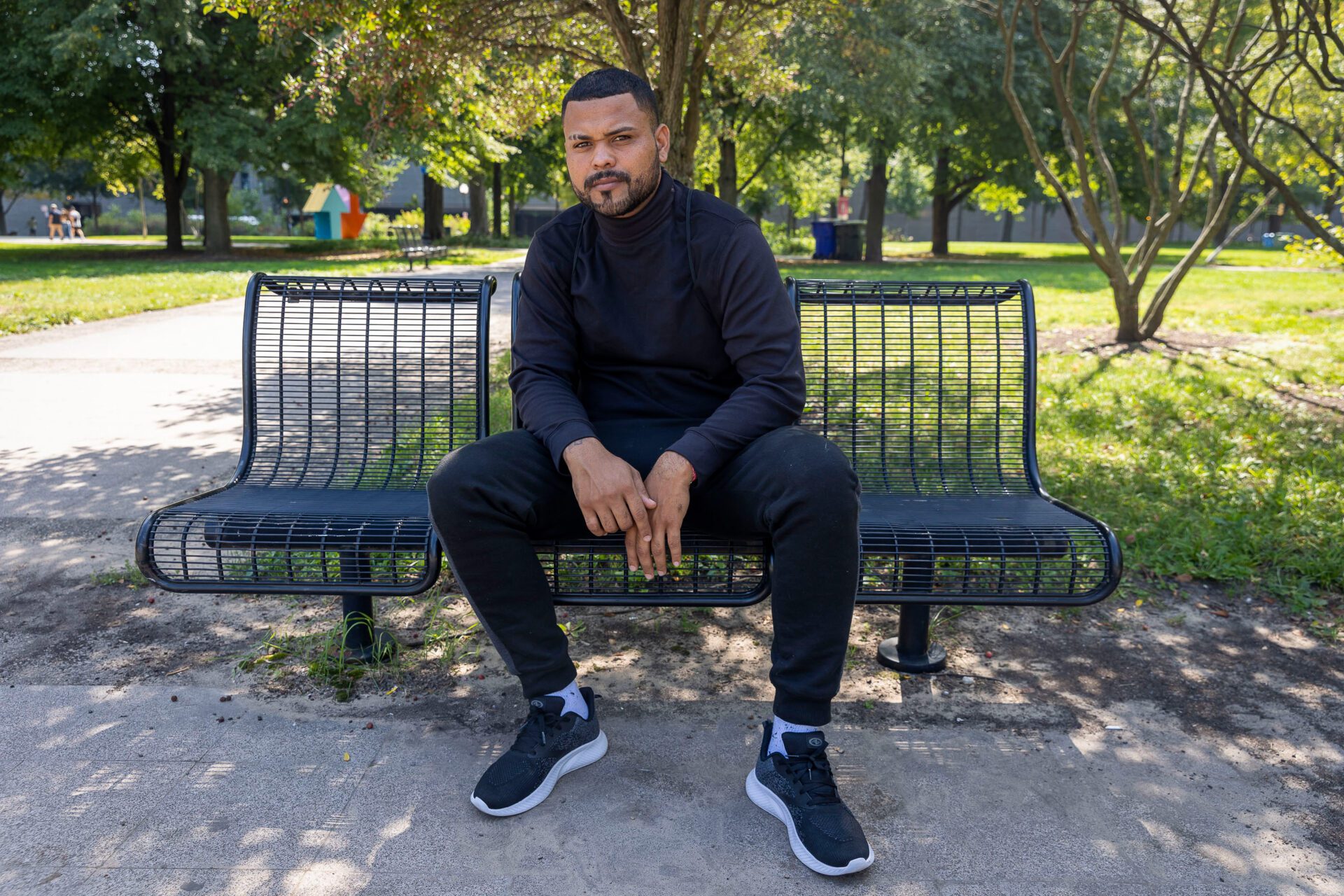 Omar con pantalón de chándal negro y cuello alto sentado en un banco del parque