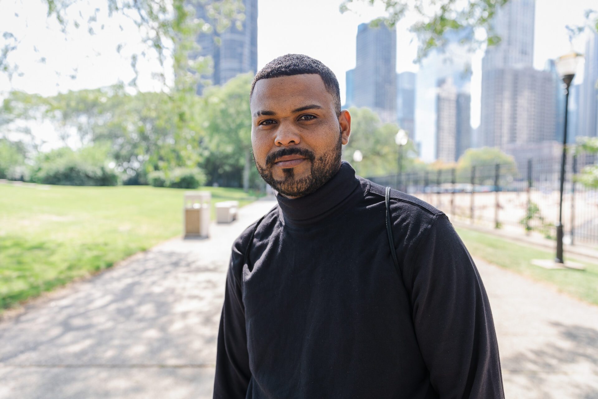 Omar with a black turtleneck and beard and mustache standing on a sidewalk, lots of skyscrapers in the background
