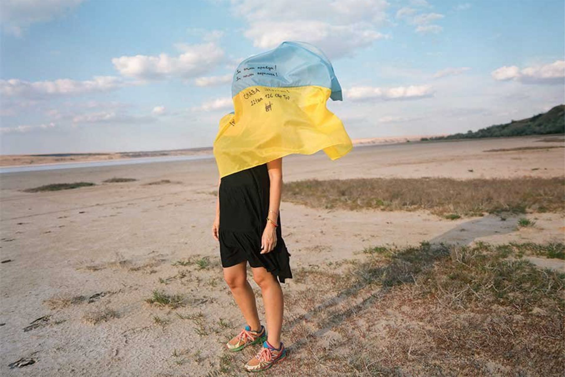 a woman on the sand and a black dress with a Ukrainian flag draped over her head and flowing in the wind