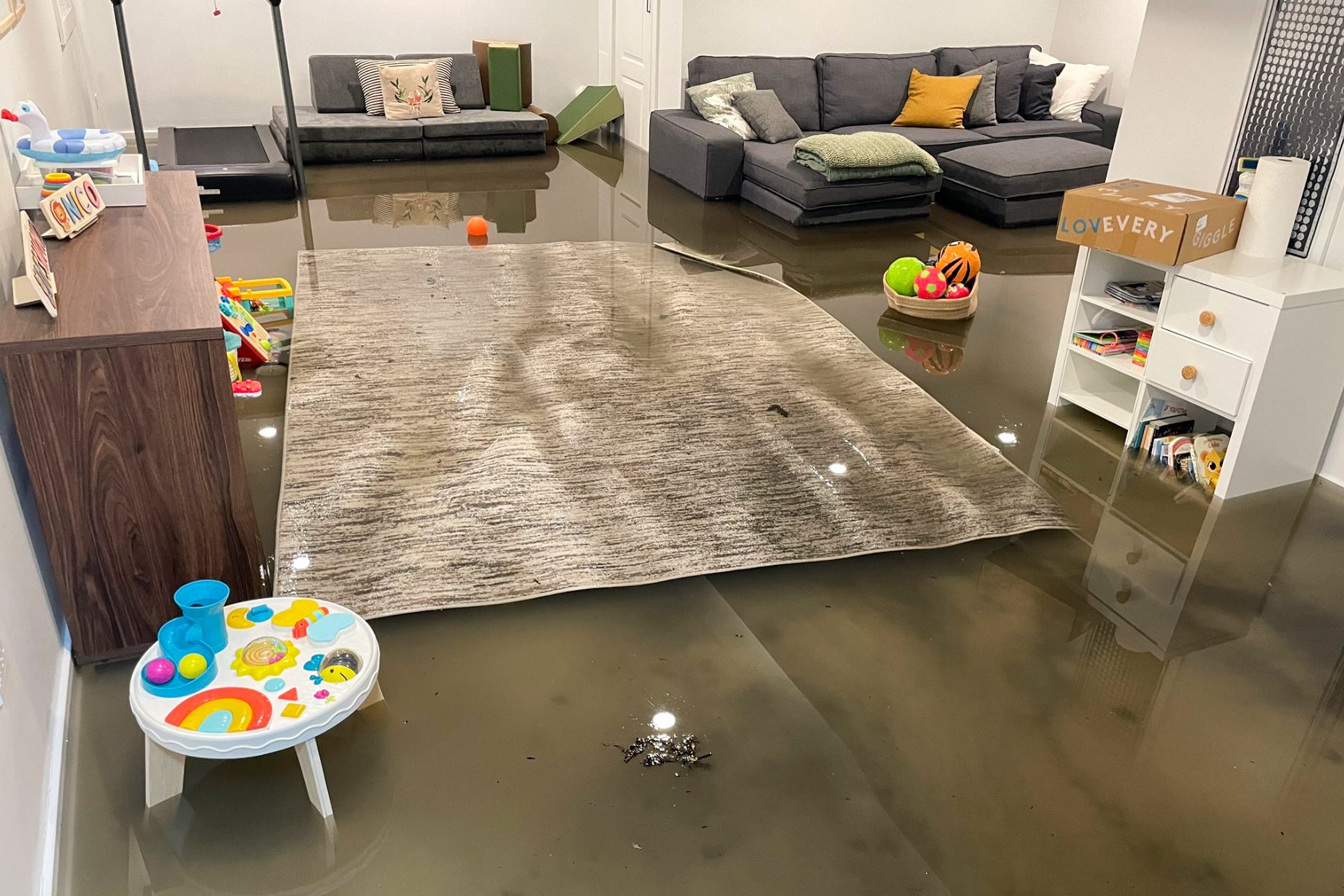 sótano inundado con varios centímetros de agua, una alfombra flotante y muebles, una cinta de correr y juguetes de bebé todo en varios centímetros de agua