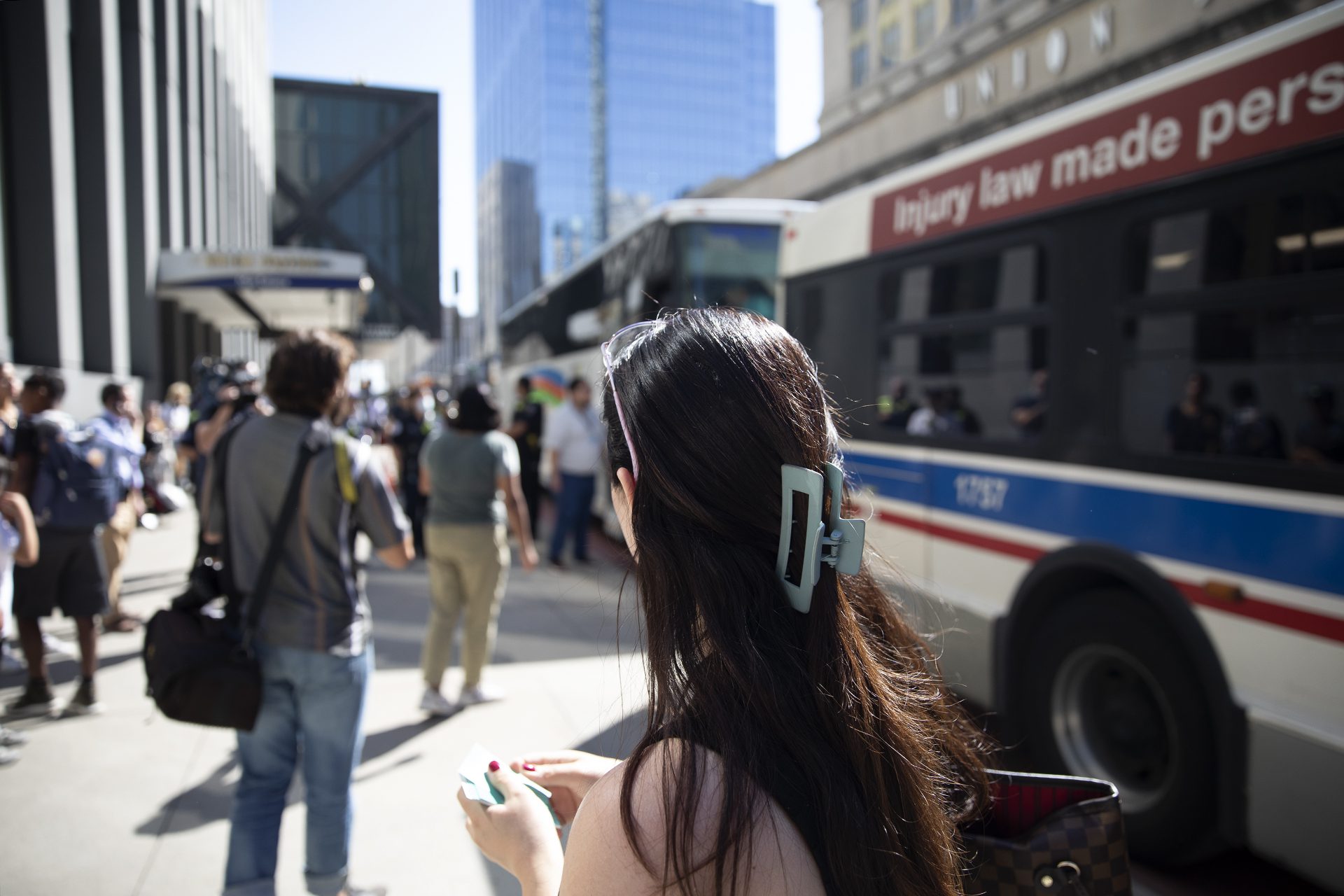 buses lined up outside Union station and migrants transferring from one to another blurry in the background; in the foreground Diane looking on with business cards in her hands