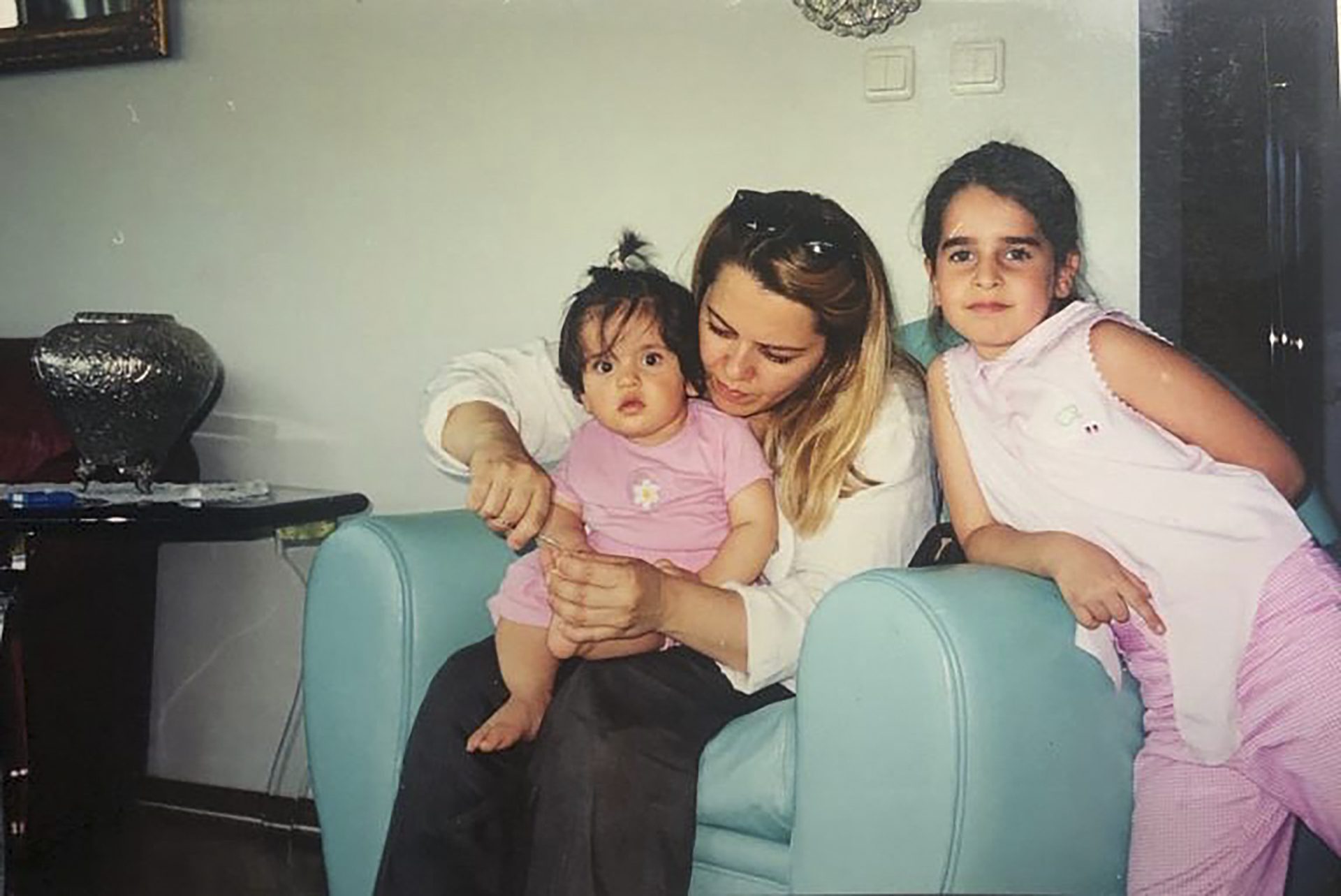 Piril de niña con su madre y su prima junto a una silla azul mientras su madre le corta las uñas de los pies a su prima pequeña.
