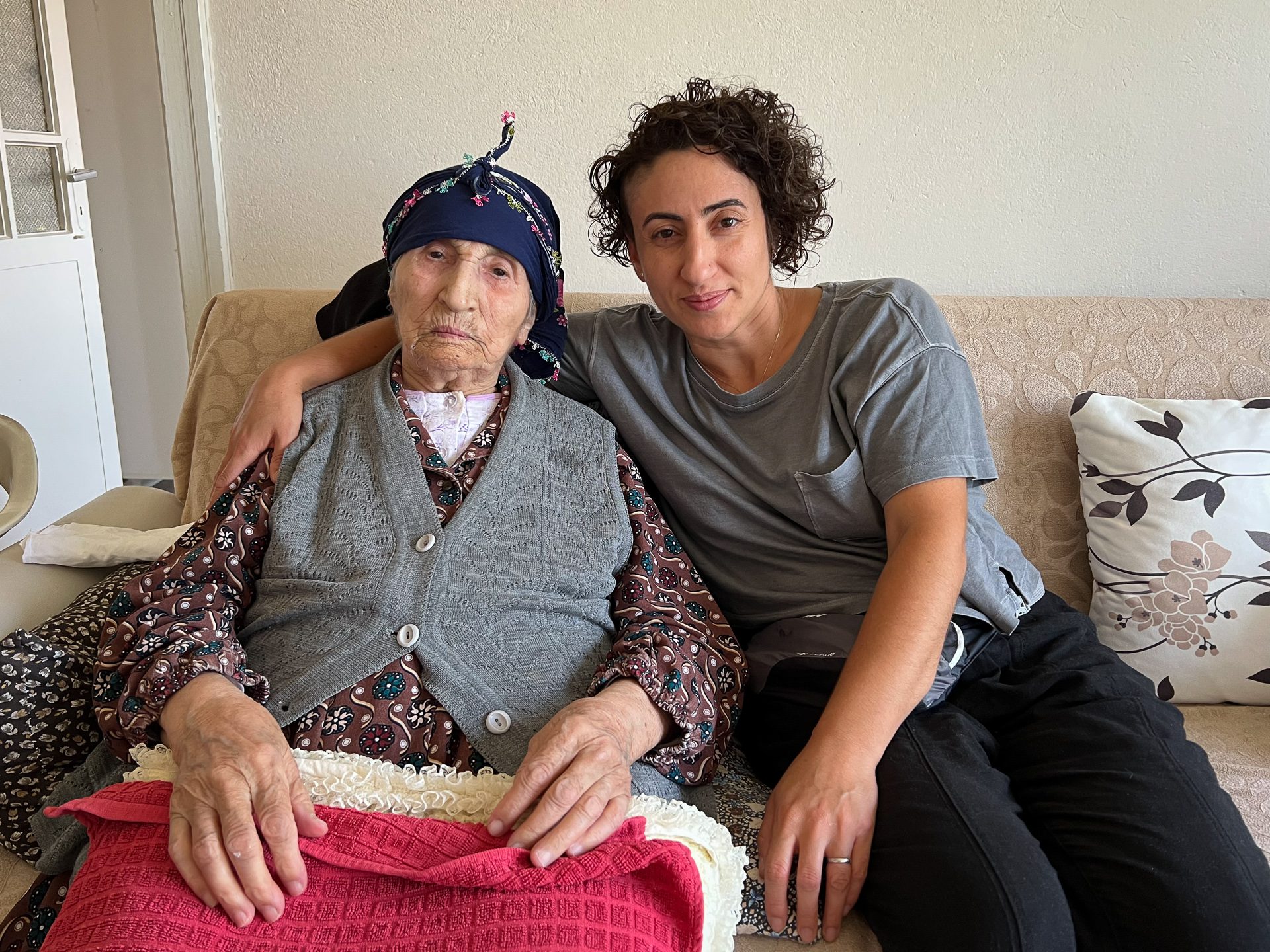 Fatma, Pinar's grandmother wearing a brown blouse with a gray cardigan and Pinar wearing black pants and gray shirt with an arm around her while sitting on a beige couch