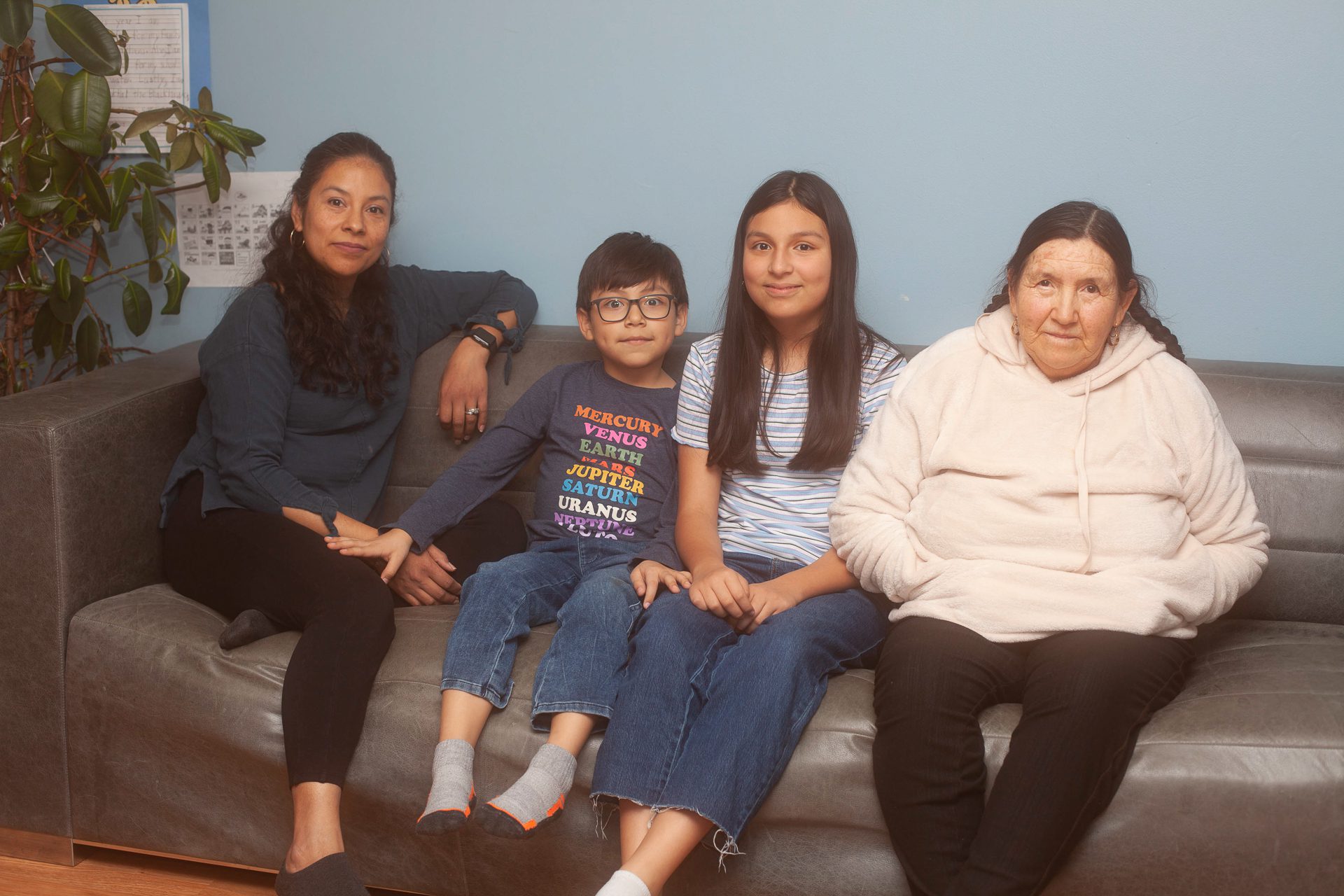 Una madre, sus dos hijos y su suegra están sentados en un sofá con una pared azul de fondo. El hijo menor tiene una camiseta con los nombres de los planetas.