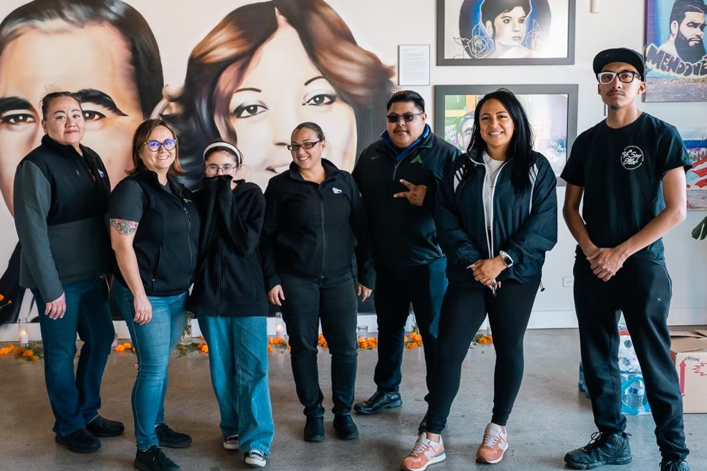 seven people stand in front of a wall filled with murals and paintings