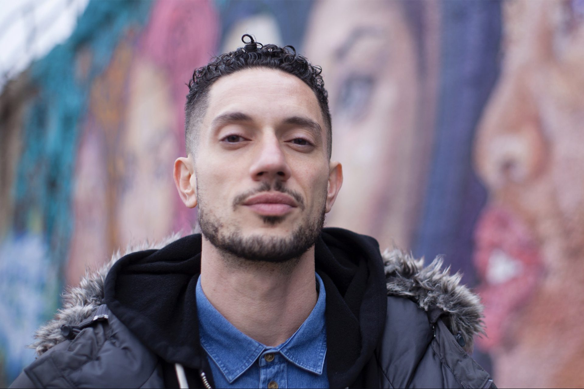 Omar Offendum wearing a jean button up, hoodie and winter coat outside in front of a mural