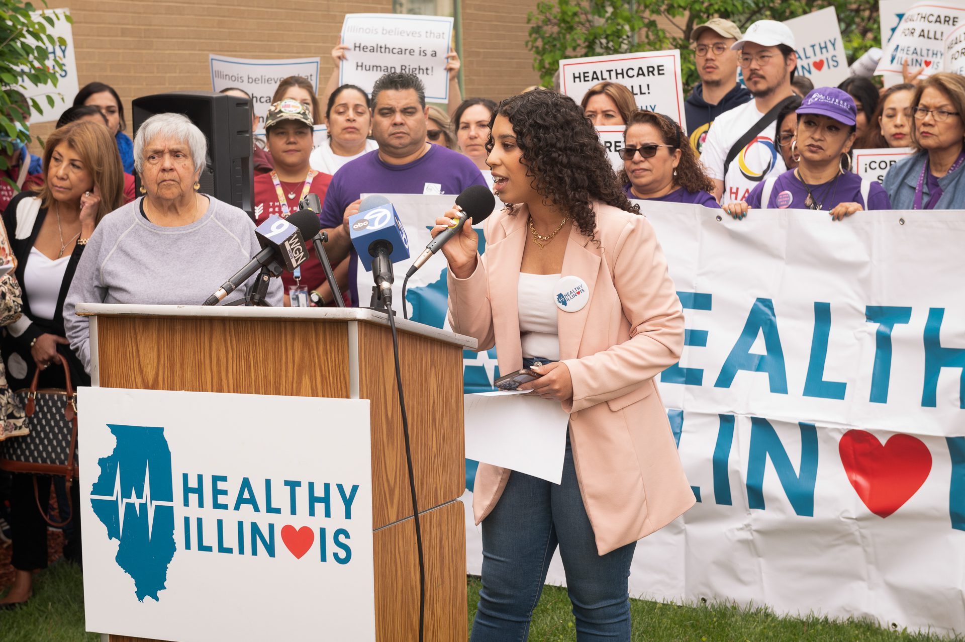 Basma Outhman addresses a crowd from behind a lectern. The lectern has a sign that reads "Healthy Illinois." Behind her is a crowd holding a "Healthy Illinois" banner and holding "Health Care for All" signs.