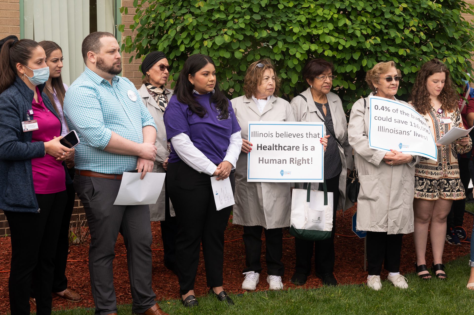 Organizers and speakers stand in the grass outside of the Alivio Medical Center in Chicago, Ill., holding signs reading "Illinois believes that health care is a human right."