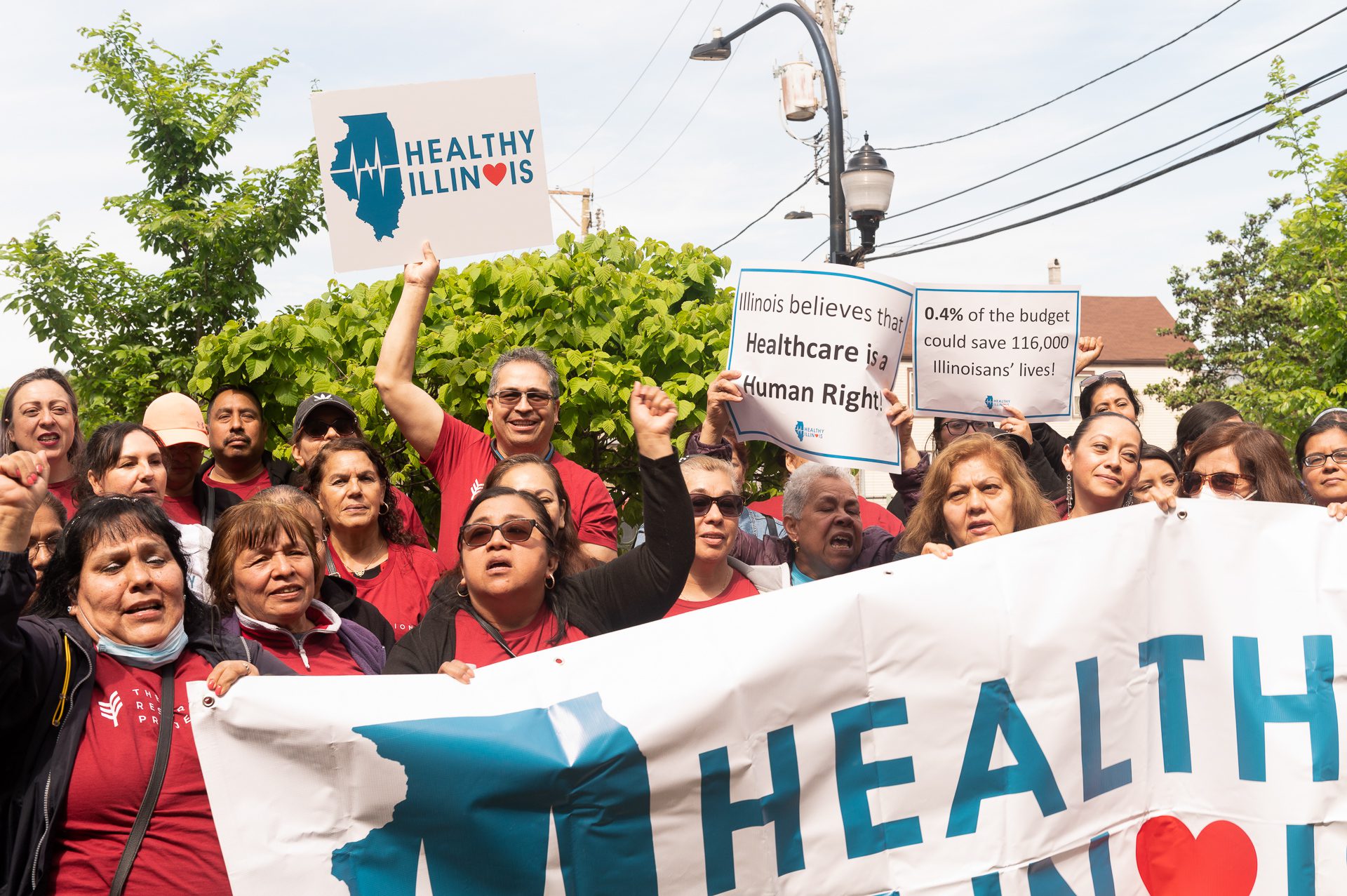 Organizadores del Proyecto Resurrección entonan cánticos en una rueda de prensa de la Campaña Illinois Saludable en el Centro Médico Alivio de Chicago (Illinois). Sostienen una pancarta y carteles en los que se lee &quot;Illinois Saludable&quot; e &quot;Illinois cree que la atención sanitaria es un derecho humano&quot;.