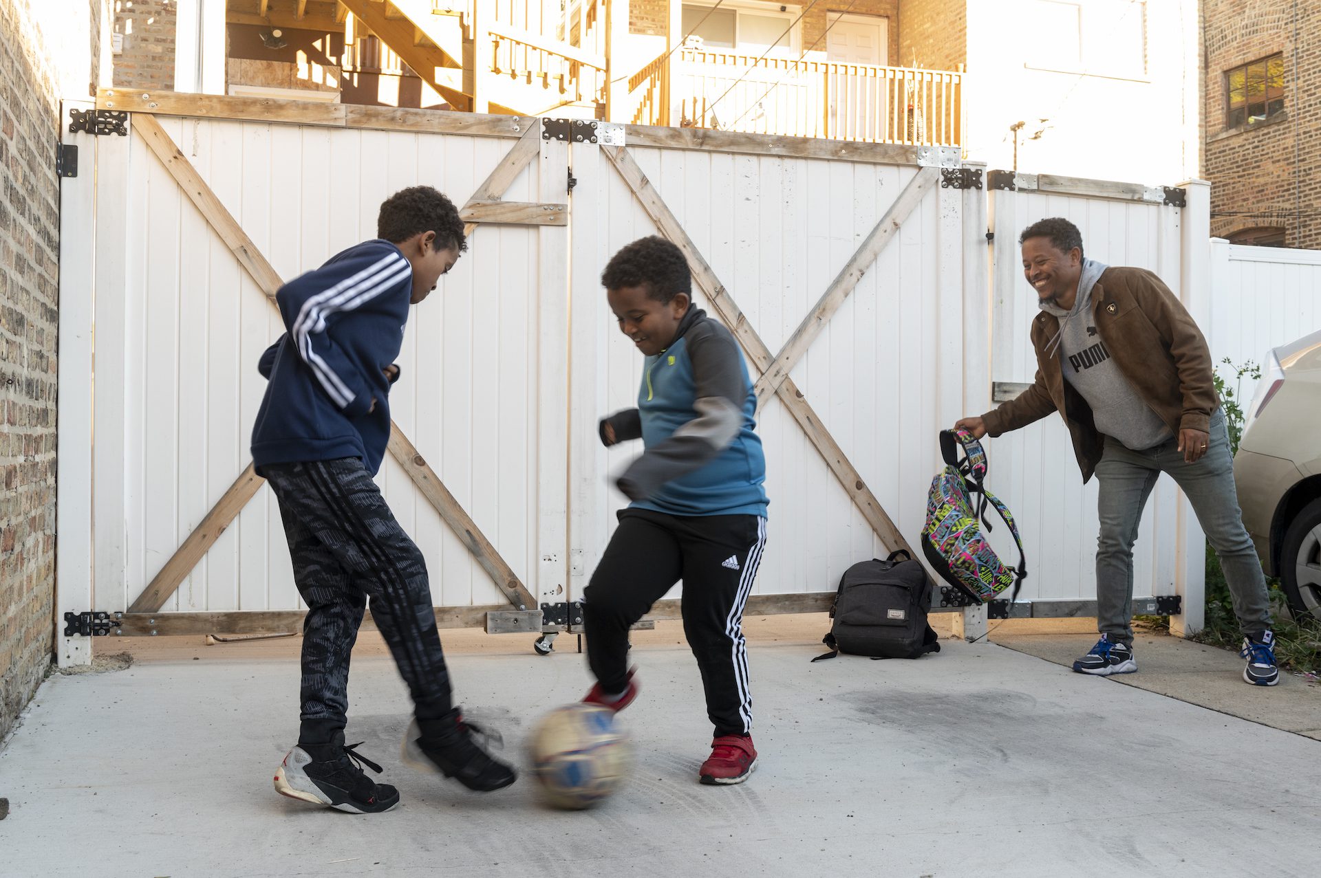 Deux enfants jouent au football tandis qu'un homme sourit en tenant un sac à dos.